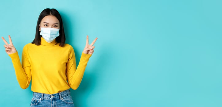 Covid-19, social distancing and pandemic concept. Kawaii asian woman showing peace signs and smiling happy, wearing medical mask from coronavirus disease, standing over blue background photo