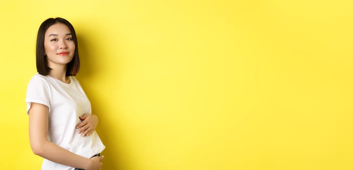 Asian woman smiling kindly, touching belly and looking at camera, being pregnant, having a baby, standing over yellow background photo