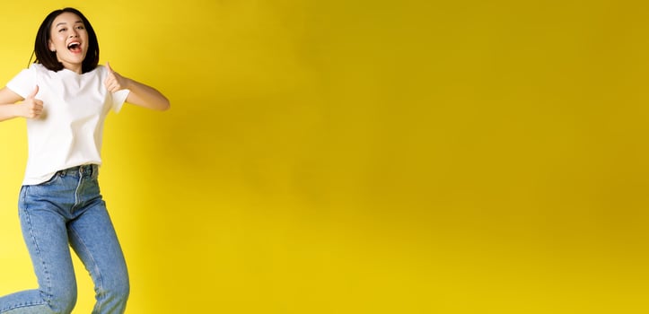 Full size of happy young asian woman jumping from joy, showing thumbs up in approval, posing over yellow background in jeans and casual white t-shirt photo