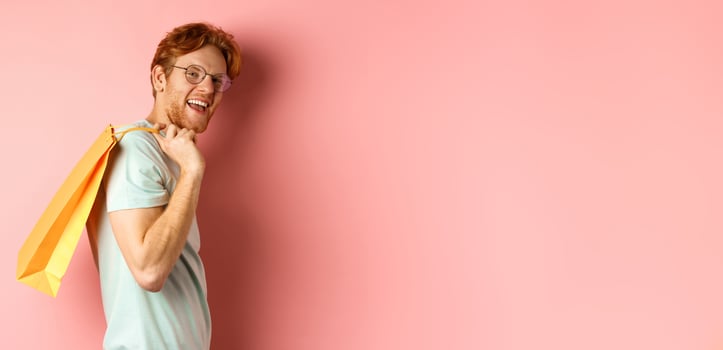 Carefree young man with red hair and glasses, walking with shopping bag over his shoulder and smiling, shopper buying presents, standing satisfied over pink background photo