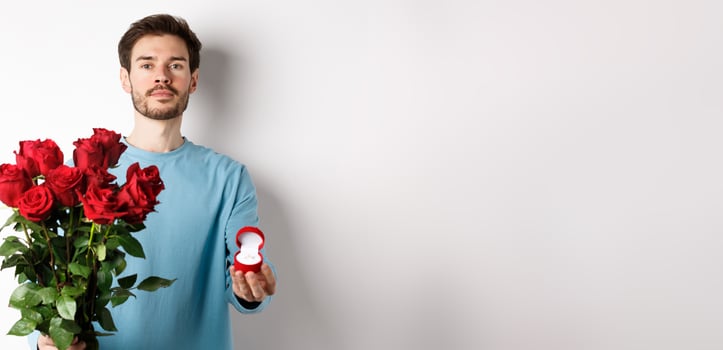 Valentines and relationship. Romantic man boyfriend holding red roses and showing engagement ring, making a proposal on lovers day, standing over white background photo