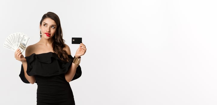 Fashion and shopping concept. Elegant woman in black dress, red lipstick, holding money and credit card, making choice, thinking, standing over white background photo