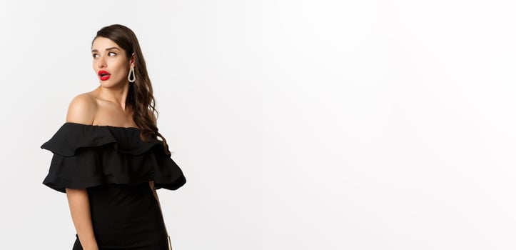 Fashion and beauty concept. Image of attractive young woman in black dress turn behind and looking at copy space, standing over white background photo