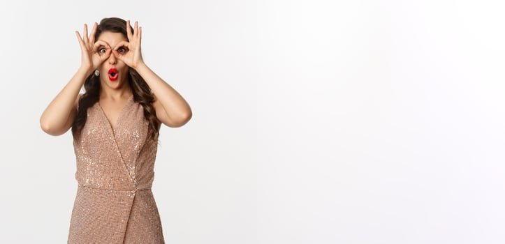 Christmas party and celebration concept. Woman in glamour dress and red lips looking at something amazing, staring through hand binoculars, standing over white background photo