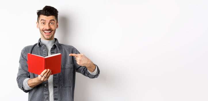 Happy smiling guy pointing at red planner, showing good journal or book, standing on white background photo