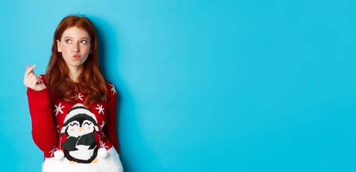 Winter holidays and Christmas Eve concept. Lovely redhead woman in xmas sweater, showing heart sign and thinking, looking upper left corner at logo, blue background photo