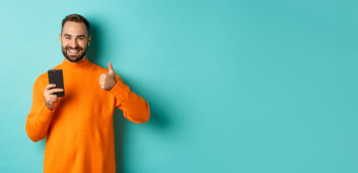 Photo of satisfied young man in orange sweater, showing thumb-up after reading on mobile phone, standing pleased against turquoise background