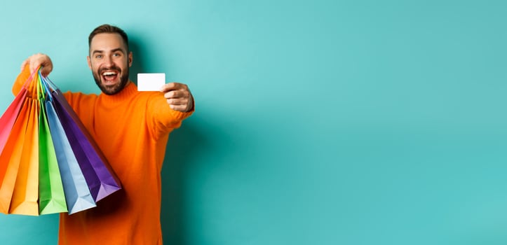 Happy aduly man showing credit card and shopping bags, standing against turquoise background photo
