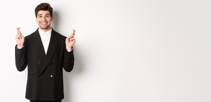 Hopeful and optimistic young businessman in suit, smiling while crossing fingers for good luck and making wish, standing over white background photo