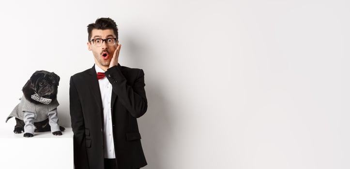 Animals, party and celebration concept. Shocked handsome man in formal suit and cute dog in costume, staring at camera amazed, standing over white background photo