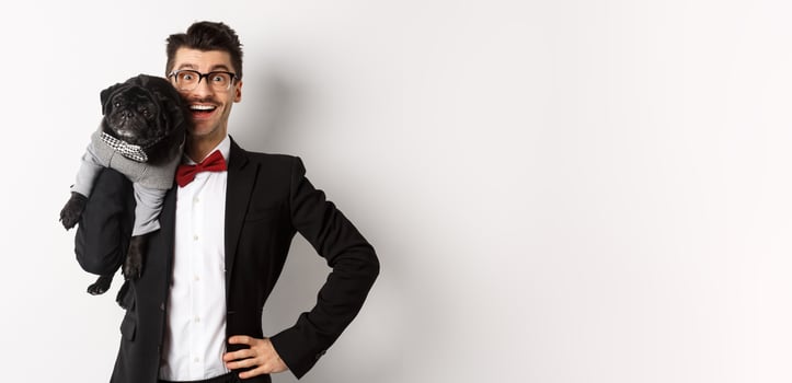 Handsome young man in suit and glasses holding cute black pug dog on shoulder, smiling happy at camera, wearing party outfits, white background photo