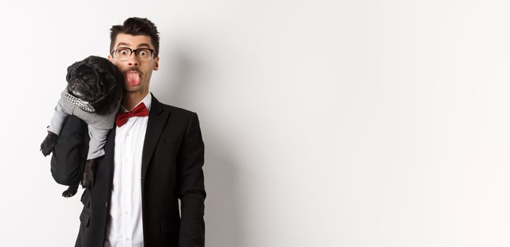 Funny young man in party suit, showing tongue and holding cute black pug on shoulder, celebrating with pet, standing over white background photo