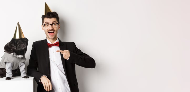 Cheerful man and cute black pug wearing party cones and suits, dog owner celebrating pet birthday, standing over white background photo