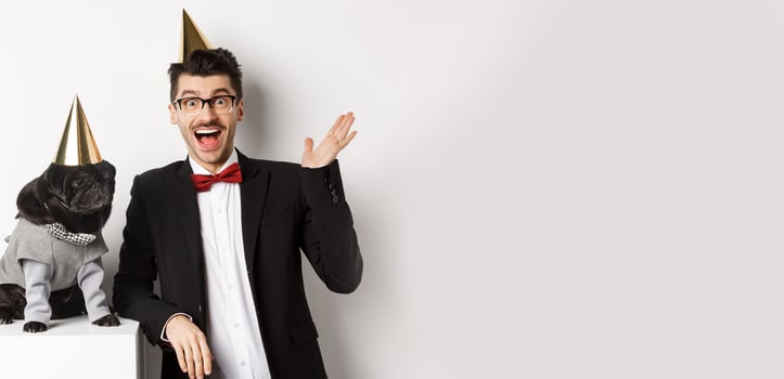 Happy young man and cute black dog wearing party cones, celebrating birthday, guy friendly saying hello and waving hand, standing over white background photo