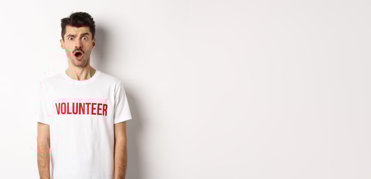Shocked and confused man in volunteer t-shirt staring at camera speechless, standing against white background photo