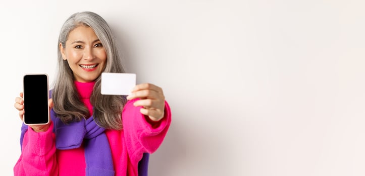 Online shopping. Close-up of stylish korean senior woman showing empty mobile screen and plastic credit card, smiling happy, standing over white background photo