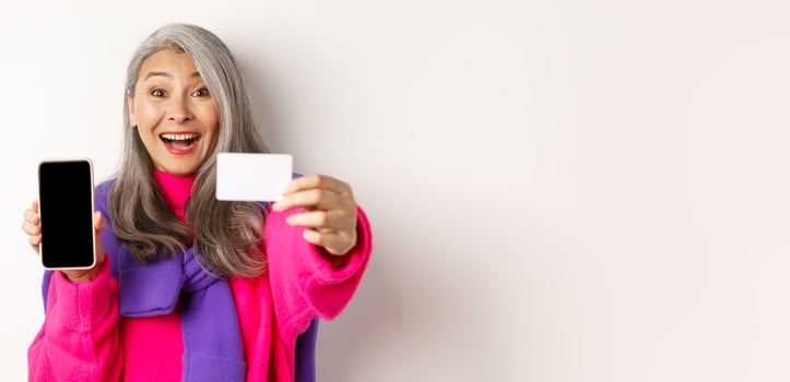 Online shopping. Close-up of stylish korean senior woman showing empty mobile screen and plastic credit card, smiling happy, standing over white background photo