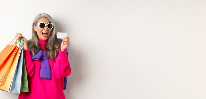 Stylish asian grandmother in sunglasses going shopping on holiday sale, holding paper bags and plastic credit card, standing over white background photo