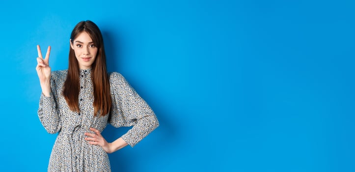 Young beautiful girl in dress with natural long hair, showing number two with fingers and smiling, standing against blue background photo
