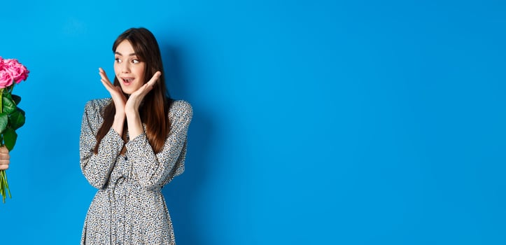 Valentines day concept. Romantic girl looking surprised at hand with flowers, woman receiving bouquet of pink roses, standing on blue background photo