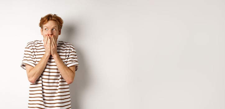 Shocked redhead guy gossiping, giggle in hands and looking left impressed, standing over white background photo