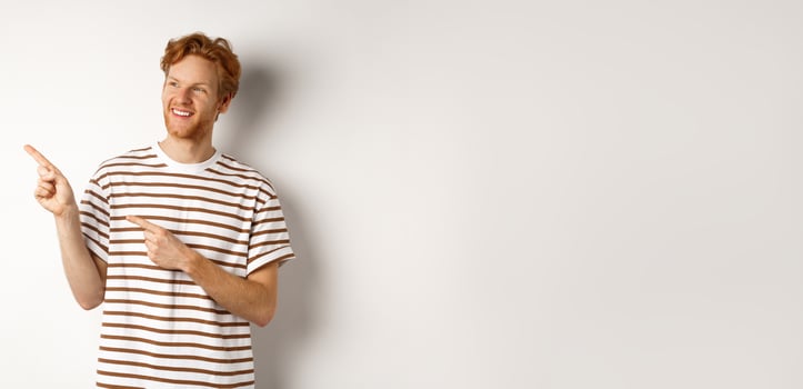 Cheerful caucasian guy with red hair and beard, looking and pointing left at logo banner, standing over white background photo
