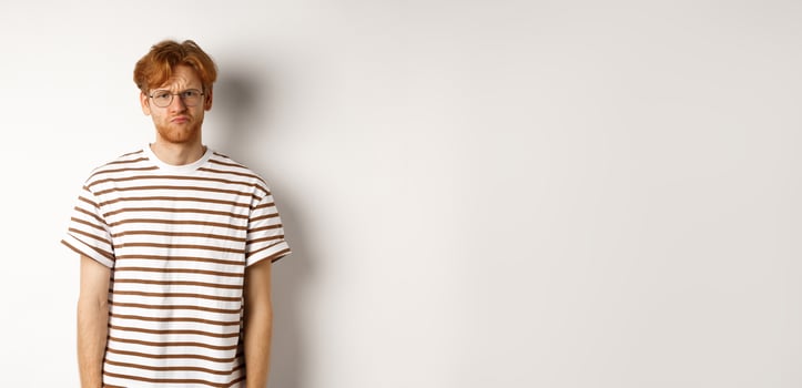 Sad and gloomy redhead male student pouting, frowning and looking distressed, standing over white background photo