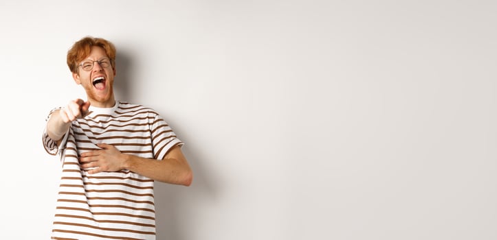 Young man with ginger hair and beard pointing finger at camera and laughing, making fun of someone hilarious, standing over white background photo