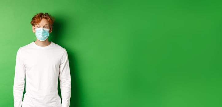 Covid-19, social distancing and lifestyle concept. Young redhead man wearing face mask during coronavirus pandemic, looking surprised at camera, green background photo