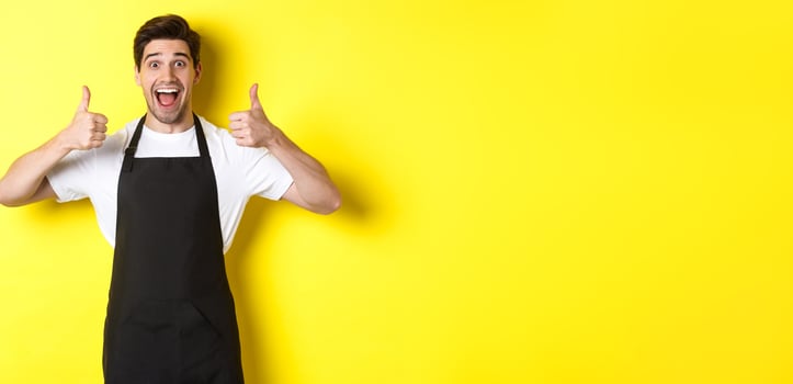 Cheerful man barista in black apron showing thumbs-up, recommending cafe or restaurant, standing against yellow background photo