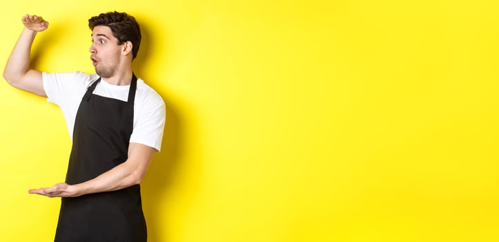 Waiter in black apron looking amazed at something large, holding big object, standing over yellow background photo