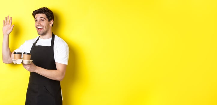 Friendly waiter in cafe waving hand at customer, holding takeaway coffee oder, standing against yellow background in black apron photo