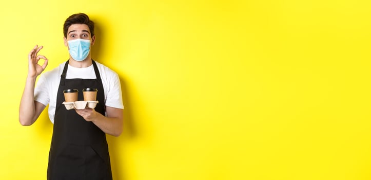 Concept of covid-19, cafe and social distancing. Barista in medical mask and black apron guarantee safety, holding takeaway cups of coffee and showing OK sign, yellow background photo