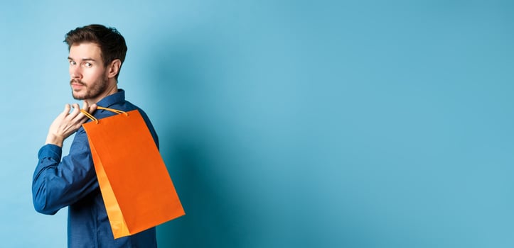 Rear view of happy man holding shopping back on shoulder and looking behind, standing on blue background photo