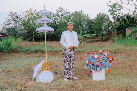 The handsome full body of a young Asian man standing among decorations wearing modern Javanese traditional clothing, a white velvet bridal kebaya in an outdoor barren garden. for beauty, fashion photo
