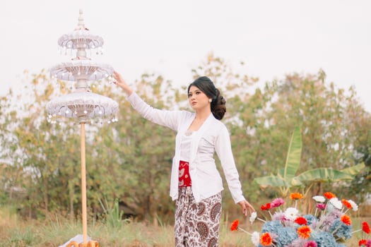 the charm of a adult Asian woman standing formally candidly holding flowers and an umbrella wearing modern Javanese traditional clothing, white velvet bridal kebaya in outdoor. for beauty, fashion photo