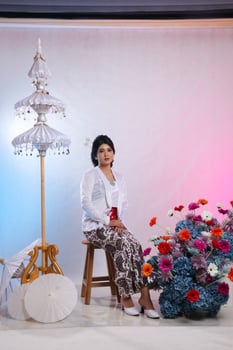Full length of young Asian girl sitting on a chair wearing modern Javanese traditional clothing, white velvet bridal kebaya on a white studio background. for advertising content, beauty, fashion photo