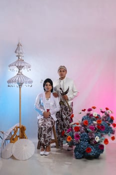 full body pair of lovers, a young Asian man and woman, sitting elegantly next to a man carrying a keris wearing modern Javanese traditional clothing, a white velvet bridal kebaya on a white background photo