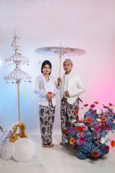 full length of a young Asian men and woman lover standing elegantly, man holding an umbrella wearing modern Javanese traditional clothing, white velvet kebaya for the bride on a white background photo