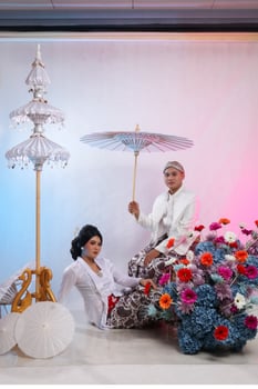 full length couple of lovers, a young Asian boy and girl, are slumped on the floor, a man sitting on a chair holding an umbrella wearing modern Javanese traditional, a white velvet bridal photo