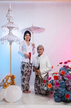 full body of a pair of young Asian male and female lovers, woman standing smiling holding an umbrella, man squatting wearing modern Javanese tradition, white velvet for the bride and groom photo