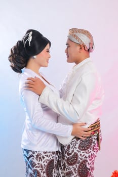 A pair of young indonesia men and woman lovers, standing smiling romantically facing each other wearing modern Javanese traditional clothing, white velvet bridal kebaya on a white studio background photo