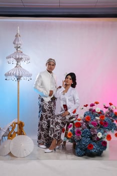 full body pair of indonesia husband and wife lovers, women smiling gracefully holding their cheeks sitting next to a man wearing modern Javanese traditional clothing, a white velvet bridal kebaya photo