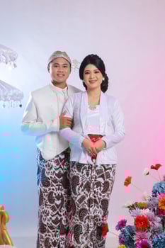 A pair of Asian lovers smiling, the man's hand holding the woman's arm, wearing modern Javanese traditional clothing, a white velvet bridal kebaya, prewedding concept on a white studio background. photo