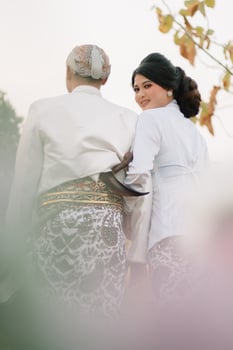 couple young Asian boy and girl walking, the woman looking at the camera holding the man wearing modern Javanese traditional clothing, the bride's white velvet kebaya in an outdoor barren garden photo
