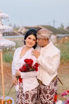 couple young Asian male and female lovers standing, smiling woman holding red flowers being caressed by man wearing modern Javanese traditional clothing, bridal white velvet kebaya.a photo