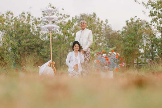 full length young Asian male and female lovers smiling, the man standing, the woman sitting below wearing modern Javanese traditional clothing, the bride's white velvet in an outdoor barren garden photo