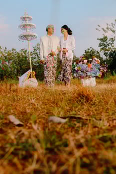 full body pair of young Asian boy and girl walking looking eye to eye amidst decorations wearing modern Javanese traditional clothing, white velvet bridal kebaya in an outdoor barren garden photo