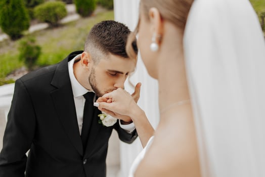 A man kisses a woman's hand in a wedding ceremony. The bride is wearing a white veil and the groom is wearing a black suit photo
