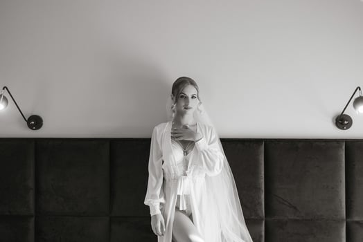 A woman in a white gown stands in front of a black headboard. She is wearing a veil and a necklace photo